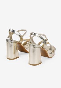 Metallisch goldene High-Heel-Sandalen mit dicken Absätzen, die mehrere übereinander gekreuzte Riemen und einen verstellbaren Schließen am Knöchel besitzen.