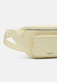 Jasnożółta nerka z miękkiego materiału. Posiada główną komorę zapinaną na zamek oraz mniejszą kieszeń na zamek z przodu. W zestawie regulowany pasek.