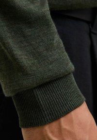 Zielony sweter dzianinowy z bliska, z prążkowanym mankietem i miękką fakturą. Tkanina pokazuje subtelne różnice w kolorze, co podkreśla jej głębię.