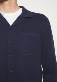 Man som bär en marinblå knappar-upp cardigan med bröstficka över en enkel vit T-shirt med rund hals.