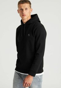 Sudadera negra con cordón en la capucha, puños acanalados y un pequeño logotipo en el pecho. Llevada sobre una camiseta blanca y combinada con jeans azul claro.