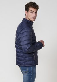 Jūrinės spalvos pūkinė striukė su siūlėmis, aukštu apykakle ir užtrauktuku priekyje. Suderinta su mėlynais džinsais. Lygi tekstūra ir lengvas dizainas.