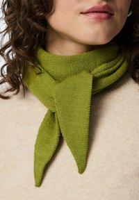 Bufanda verde de punto con un acabado texturizado, con un diseño atado y puntas puntiagudas. Llevada sobre una prenda de un tono beige claro.