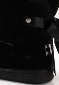 Botín de invierno de ante negro con forro de piel de borrego, lazo de cinta, placa de logo de metal y detalle de botón de metal de Mustang Shoes en el talón.