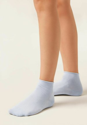 Persona che indossa calzini alla caviglia azzurri chiaro, in piedi su una superficie neutra con i piedi piatti e le gambe visibili fino a metà polpaccio.