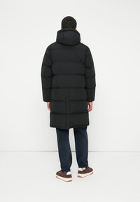Mand iført en lang sort polstret frakke med hætte, mørkeblå jeans og brune sneakers, stående overfor en hvid væg.