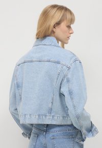 Donna bionda vista di spalle che indossa un giubbotto di jeans corto azzurro e jeans a vita alta coordinati su uno sfondo bianco semplice.