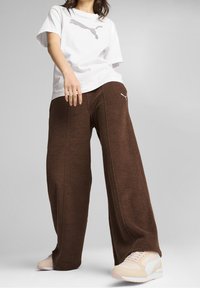 Žena v bílém tričku Puma, hnědých širokých kalhotách a béžových teniskách Puma stojící na jednobarevném šedém pozadí.