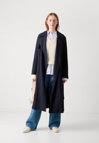 Abrigo trinchera navy con puños de piel sintética sobre un suéter crema y una camisa a rayas azules, combinado con jeans de corte relajado y zapatillas de plataforma blancas.