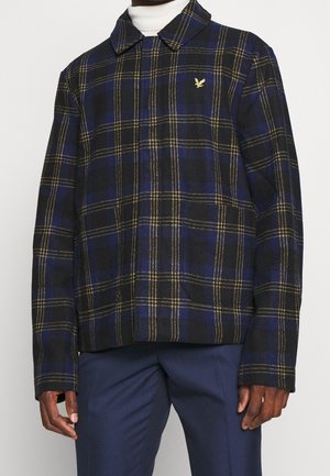 Mand iført sort og blå ternet jakke med gule linjer og gyldent fuglelogo, kombineret med marineblå bukser og hvid rullekrave.