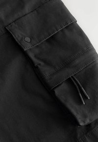 Poche de pantalon cargo en tissu noir avec rabat à bouton-pression et coutures visibles, dotée de cordons de serrage noirs.