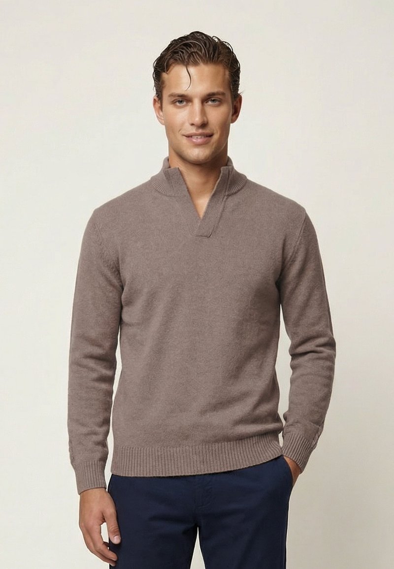 Uomo in piedi davanti a uno sfondo semplice che indossa un maglione taupe con collo a V e pantaloni blu navy.