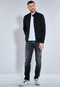 Black bomber jacket with a standing collar, zip closure, and long sleeves. Worn over a white t-shirt, paired with slim-fit gray jeans and white sneakers.