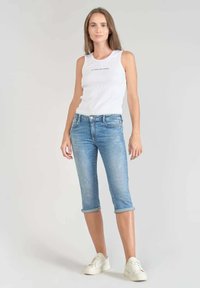 Débardeur blanc à côtes avec un détail imprimé, associé à un jean en denim bleu clair coupé et retroussé au bas, et des baskets blanches.