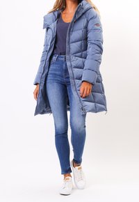 Ljusblå pufferjacka med huva, med quiltat mönster, långa ärmar och dragkedja. Kombinerad med tajta blå jeans och vita sneakers.