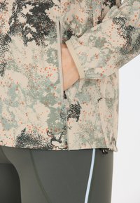 Takki, jossa on camo-kuvio beige-, vihreä- ja oranssiväreillä; siinä on vetoketjullinen tasku ja teksturoitu kangas; yhdistetty vihreisiin legginsseihin.
