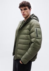 Νεαρός άνδρας που φοράει μπουφάν σε ελαιόχρωμο με κουκούλα και φερμουάρ στην τσέπη του μανικιού με ετικέτα «BOSS», με τα χέρια στις τσέπες μαύρου παντελονιού, ουδέτερο φόντο.