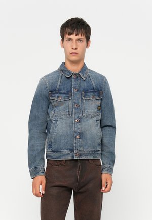 Jonge man in een vervaagde blauwe denimjack met knoopsluiting en donkerbruine broek, staand tegen een effen witte achtergrond.