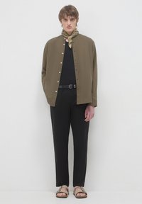 Camicia a bottoni oliva, maglietta nera, pantaloni neri, sandali marroni e sciarpa a motivi intorno al collo. Design minimalista con texture sottile.