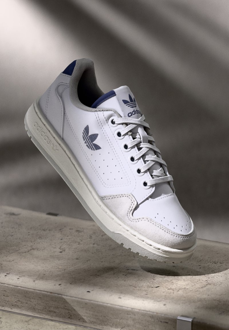 Zapatilla deportiva blanca con parte superior de cuero liso, detalles en gamuza gris y talón azul marino. Presenta perforaciones y cordones planos.