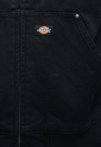 Κοντινό πλάνο τσέπης μαύρου υφασμάτινου σακακιού με μπαλώματα λογοτύπου Dickies σε πορτοκαλί, κίτρινο και μπλε, ραμμένα πάνω από το φερμουάρ.