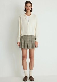White zip-up sweater with a textured finish, paired with a plaid pleated skirt. Brown loafers and cream socks complete the outfit.