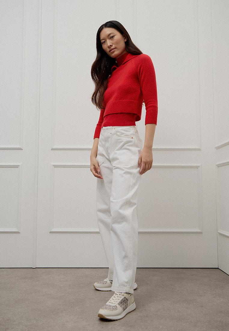 Roter, kurzer Pullover, weiße High-Waist-Jeans und helle Sneaker mit strukturiertem Muster. Neutraler Hintergrund und geradlinige Pose.