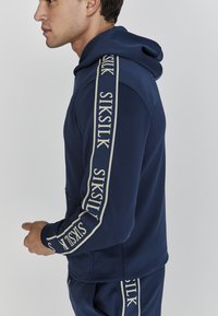 Felpa navy con tasca frontale e cappuccio regolabile con cordino. Presenta il logo beige "SIKSILK" lungo le maniche. Tessuto morbido e elastico.