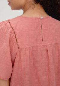 Pfirsichfarbene Bluse aus einem strukturierten Stoff, mit einem gefalteten Rücken und einer kleinen Schlitzöffnung, die am Ausschnitt mit einem Knopf gesichert ist.