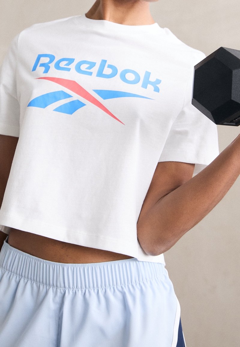 Bílý zkrácený tričko s modrým a červeným logem Reebok. Osoba drží černou šestihrannou činku, v kombinaci se světle modrými šortkami.