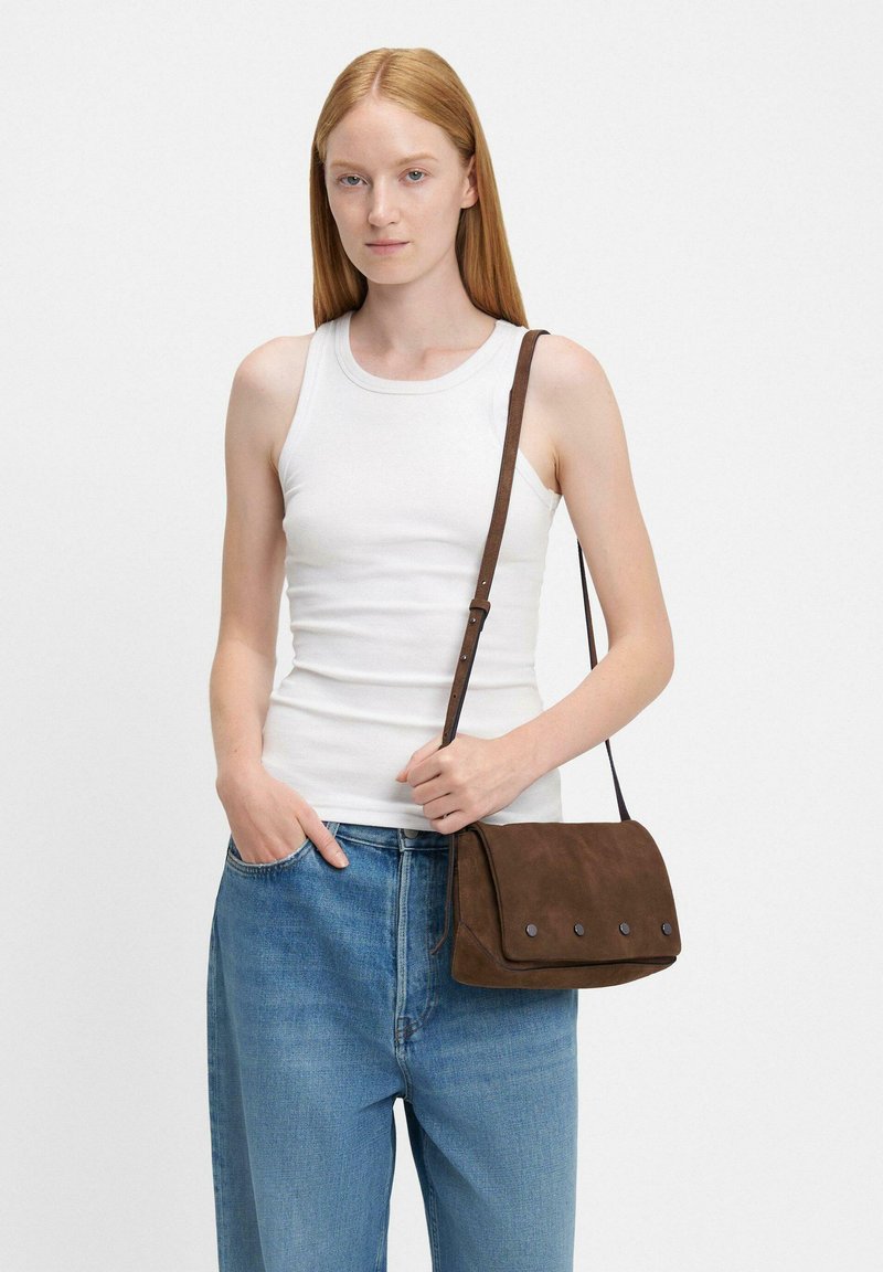 Donna in top senza maniche bianco e jeans blu che tiene una borsa a tracolla in suede marrone con borchie metalliche su sfondo uniforme.