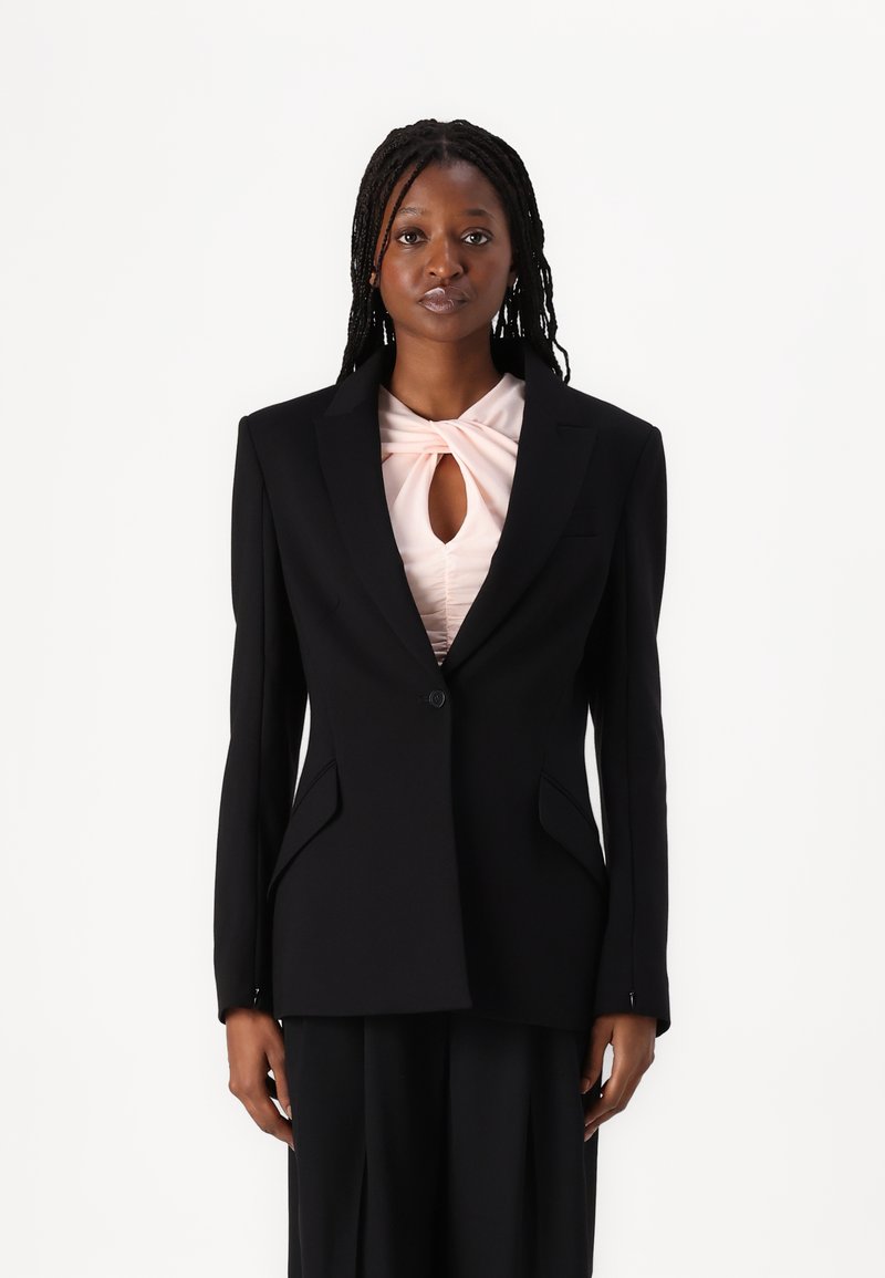 Blazer nera su misura con rever affilati, due tasche frontali e chiusura con un solo bottone, indossato sopra un top rosa plissè con dettaglio a chiave.