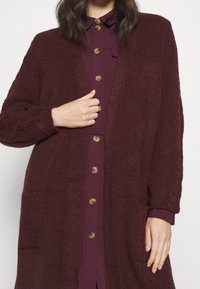 Burgundowy kardigan o teksturowanym splocie, z otwartym przodem i dwoma kieszeniami, noszony na koszuli z guzikami i kołnierzykiem w ciemniejszym odcieniu burgundu.
