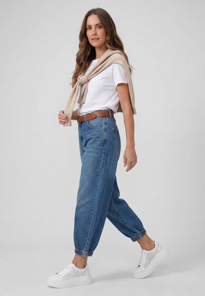 Vrouw loopt, draagt witte sneakers, blauwe omslagjeans, wit T-shirt, bruine riem en een beige trui over de schouders.