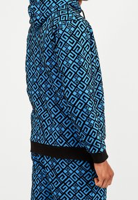 Modrá mikina s kapucí s geometrickým vzorem a černými prvky. Vyrobena z látky s hladkým povrchem, obsahuje kapuci a žebrovaný lem.