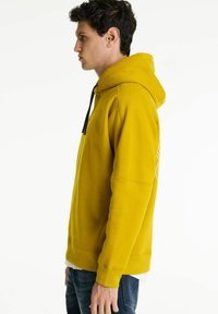 Ung mand i gul hættebluse med sorte snore, stående i profil mod en ensfarvet hvid baggrund.