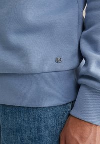 Gaiši zils džemperis, kas izgatavots no mīksta kokvilnas, ar ribotu piedurkņu un apakšmalu, ar mazu sudraba logotipa akcentu tuvu apakšai.