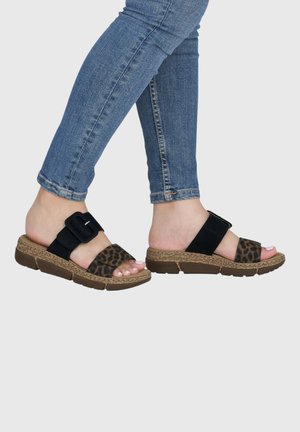 Füße in blauen Skinny-Jeans, die Slip-On-Sandalen mit schwarzen Riemen und leo-Print-Frontbändern auf braunen, strukturierten Sohlen tragen.