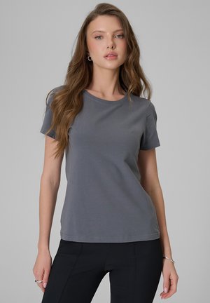 Młoda kobieta z długimi falującymi włosami, ubrana w szary t-shirt z krótkim rękawem i czarne spodnie na jednolitym szarym tle.