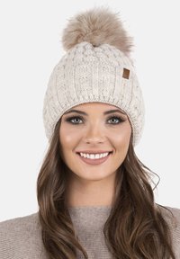 Gorro de lana blanca con un patrón texturizado, que presenta un gran pompón de piel sintética marrón claro en la parte superior y una pequeña etiqueta de cuero marrón.