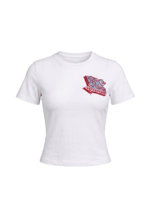 Femme aux cheveux noirs courts portant un t-shirt court blanc et un jean flare taille haute bleu clair, posant devant un fond gris.