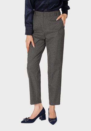 Mujer con pantalones grises a medida, blusa azul marino y zapatos azul marino con tacón bloque y detalle de hebilla cuadrada, posando frente a un fondo liso.