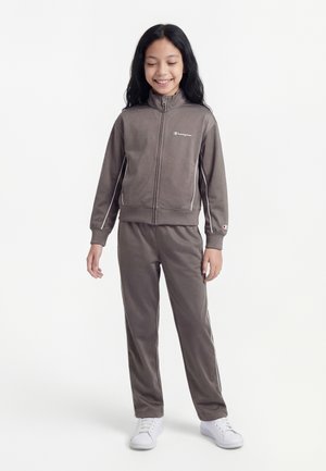 Copil cu păr negru lung, zâmbind, purtând o jachetă gri cu fermoar și pantaloni asortați, cu adidași albi, pe un fundal simplu.