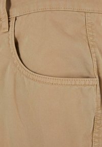 Tissu en coton beige avec une poche présentant des détails de couture visibles et une texture lisse. La surface est sans pli et uniforme.