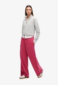 Felpa grigia con zip indossata sopra una camicia bianca, abbinata a pantaloni della tuta rosa larghi e sneaker bianche con dettagli scuri.