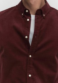 Bordeauxkleurige corduroy blouse met kraag en witte knopen, open gedragen over een witte crewneck t-shirt, waarbij de nek en bovenste borst zichtbaar zijn.