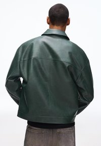 Mand vendt væk iført en mørkegrøn oversized læderjakke og falmede grå jeans mod en ensfarvet hvid baggrund.