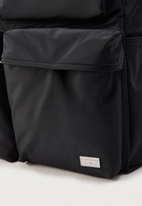 Svart nylonsekk med flere lommer, jevn tekstur, horisontale sømmer og en metallisk logo foran.