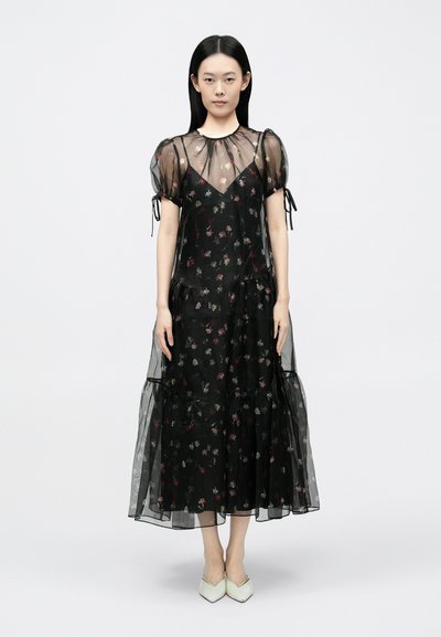 Schwarzes Blumenmuster-Kleid mit Puffärmeln, einem Lagen-Design und transparentem Stoff; mit Kordelzug-Akzenten und einem fließenden Saum. Kombiniert mit hellen Schuhen.