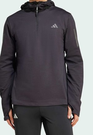 Personne portant un haut de sport noir à manches longues avec une fermeture éclair au quart et des trous pour les pouces, associée à un legging noir, debout devant un fond uni.